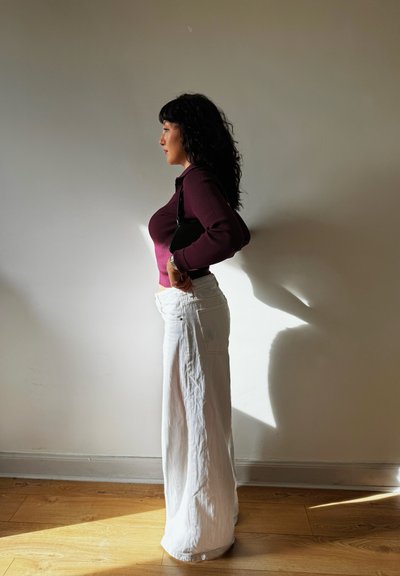 Figura curvilínea mostrada de perfil, vistiendo una blusa de manga larga burdeos con relieves y un pantalón blanco ancho, sosteniendo un pequeño bolso negro.