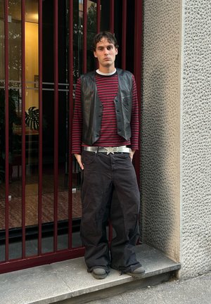 Chaleco de cuero negro sobre una camisa de manga larga a rayas rojas y negras, pantalones negros holgados, hebilla de cinturón en forma de estrella plateada, zapatos oscuros.