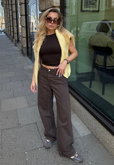 Top corto negro, chaleco de punto amarillo, pantalones anchos marrones, zapatillas grises con detalles blancos. Los accesorios incluyen gafas de sol de gran tamaño y un reloj.