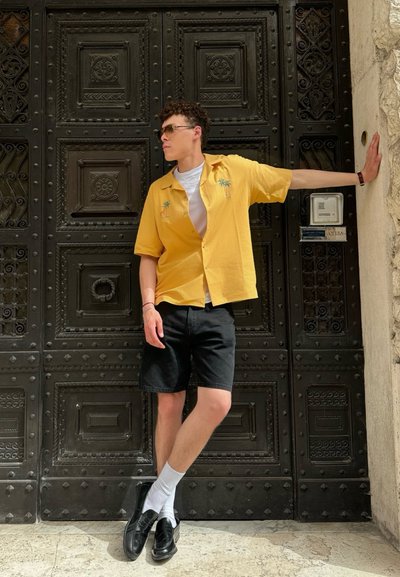 Camisa amarilla de manga corta con bordado de palmera, combinada con pantalones cortos negros. Zapatos negros y calcetines blancos. De pie frente a una puerta negra texturada.