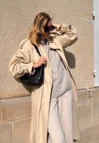 béžový trenčkot přes světle šedý svetr, spárovaný s krémovými kalhotami. Černá kabelka přes rameno a sluneční brýle dokončují vzhled. Hladké textury.