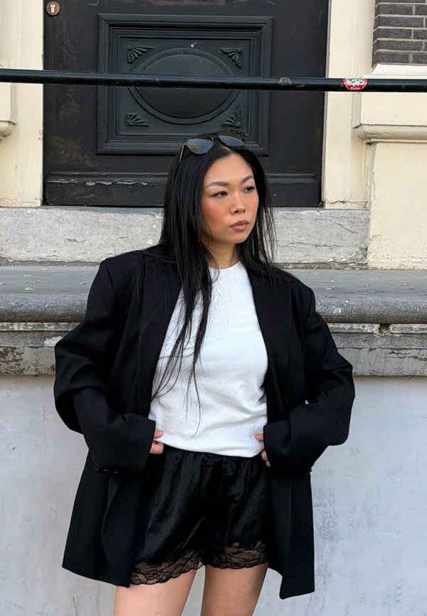 Frau mit langen schwarzen Haaren, die eine Sonnenbrille auf dem Kopf trägt, weißes Hemd, schwarzen übergroßen Blazer und schwarze Satinshorts mit Spitzenbesatz, stehend an Steintreppen.