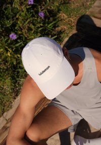 Biała czapka bejsbolowa z czarnym logo "lululemon", gładki materiał, zaokrąglona korona i zakrzywiony daszek, na tle zielonego liścia.