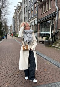 Beige long coat over grey scarf, black patterned wide-leg trousers, white shoes, and a brown bag with a white logo on a cobbled street.