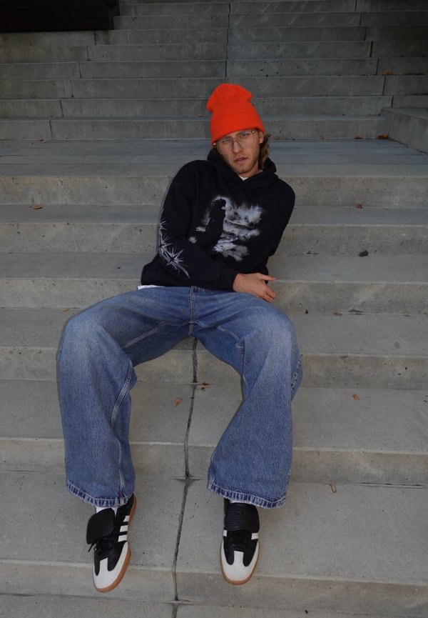 Sweat à capuche noir avec imprimé graphique, jean large bleu, et baskets noires et blanches. Un bonnet orange vif ajoute une touche de couleur. Assis sur des escaliers.