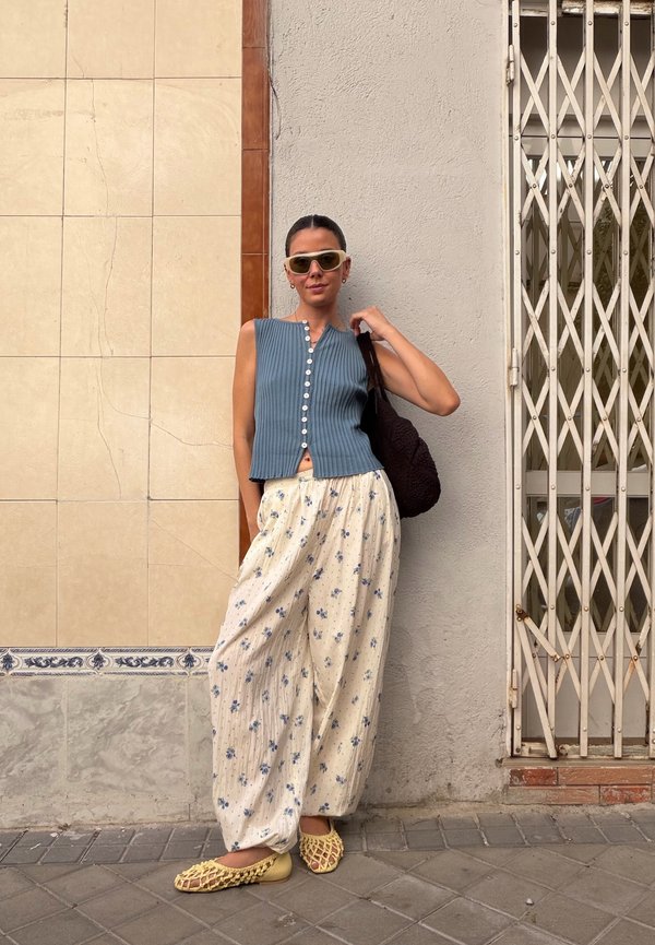 Donna con top blu senza maniche con bottoni, pantaloni bianchi larghi con stampa floreale, scarpe beige intrecciate e grandi occhiali da sole, appoggiata a un muro vicino a un cancello di metallo.