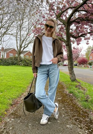 Mujer con gafas de sol, camisa blanca, chaqueta a cuadros, vaqueros y zapatillas de deporte está de pie en la acera bajo cerezos en flor, sosteniendo una bolsa negra.