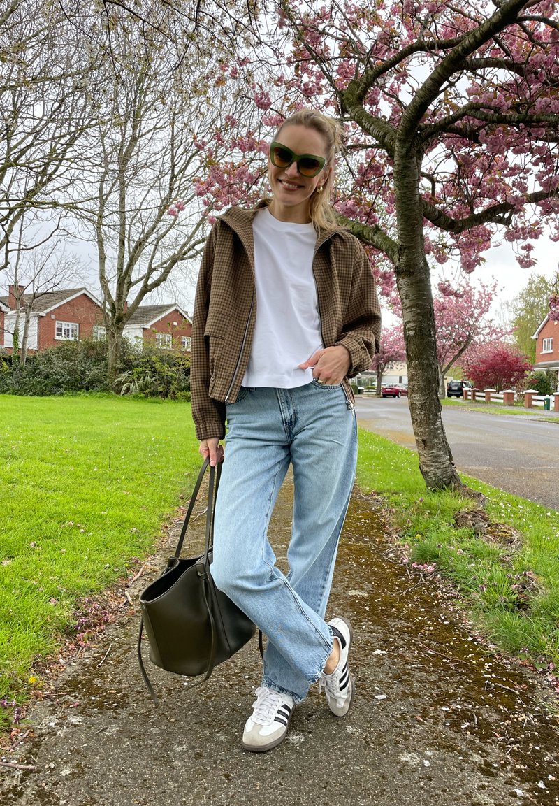 Femme portant des lunettes de soleil, une chemise blanche, une veste à carreaux, un jean et des baskets, debout sur le trottoir sous des cerisiers en fleurs, tenant un sac noir.