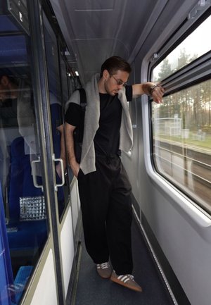 Une personne se tient dans un couloir de train, portant une chemise noire, un pull gris drapé sur les épaules, un pantalon noir et des chaussures marron avec des rayures blanches.