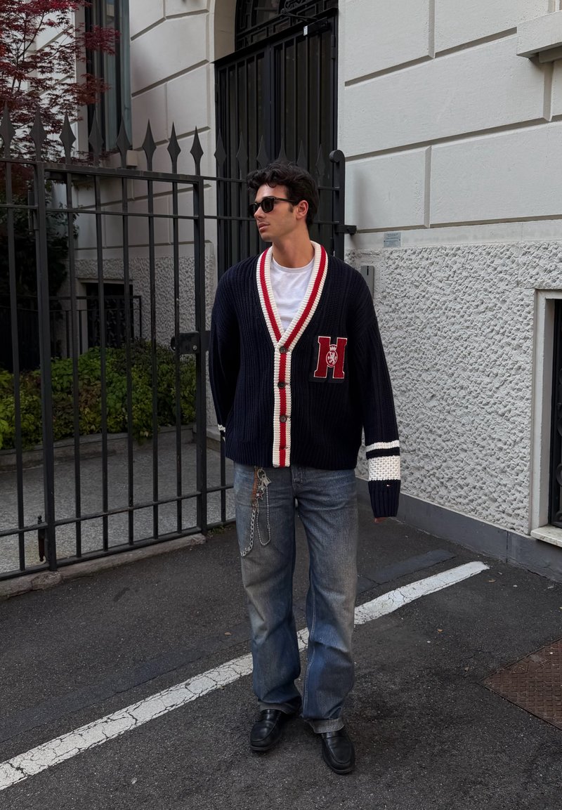 Homme portant des lunettes de soleil, cardigan bleu marine avec bordure rouge et blanche, t-shirt blanc, jeans avec chaîne, debout dans la rue près d’un bâtiment clos.