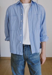 Ljust blå button-up skjorta med vita och navy vertikala ränder, med två bröstfickor, uppvikta ärmar, buren över en vit T-shirt och jeans.