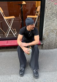 Crna majica, crna kapa i labave crne hlače. Drži prozirnu čašu s drvenom slamkom, sjedi na crvenoj klupi uz kamen zid.