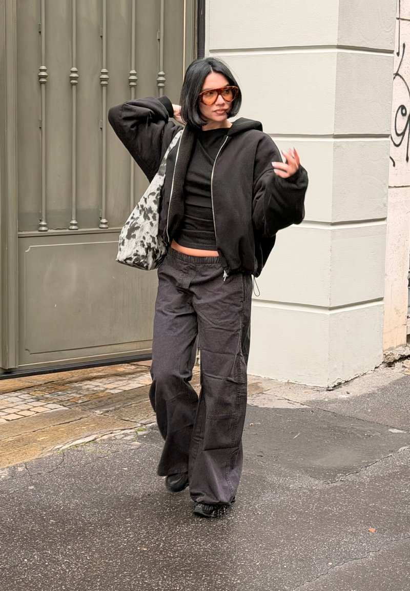 Casaco bomber preto com fecho de correr, top preto justo, calças cargo negras soltas, óculos de sol oversized e uma mala de ombro cinza e preta com padrão.