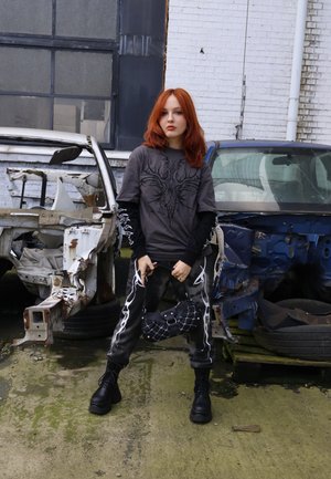 Mujer joven de cabello rojo de pie entre dos carrocerías de coches descapotados, vistiendo pantalones negros con gráficos, capas de blusas y sosteniendo un bolso acolchado.