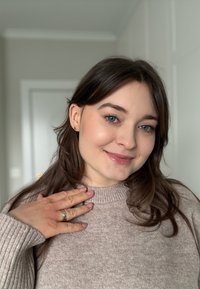 Jeune femme aux longs cheveux bruns et aux yeux bleus sourit, portant un pull beige, montrant une bague sur sa main gauche reposant près de son épaule.