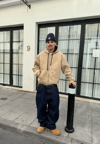 Sudadera con capucha beige, vaqueros oscuros y holgados, botas color piedra. De pie junto a un poste negro sobre un pavimento gris, con un fondo de edificio blanco.