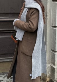 Casaco de lã castanho com grandes bolsos, combinado com um cachecol cinza claro com nervuras. O casaco tem um corte descontraído e o cachecol cai de forma solta.