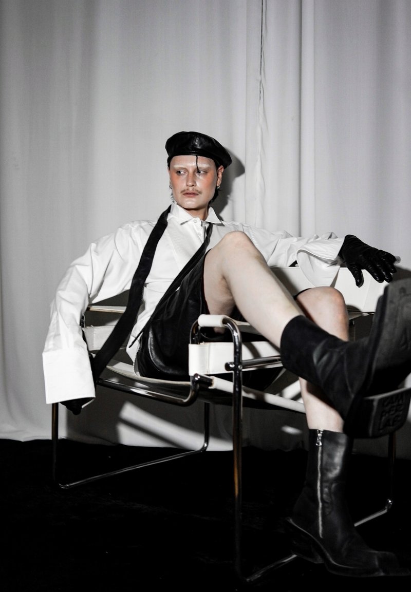 Άτομο φορώντας μαύρο δερμάτινο μπερέ, γάντια, μπότες, λευκό φαρδύ πουκάμισο και μαύρα δερμάτινα σορτς, καθισμένο με σταυρωμένα πόδια σε καρέκλα με μεταλλικό πλαίσιο.