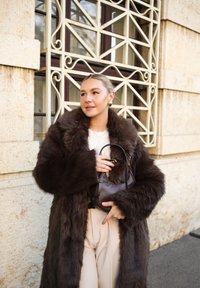 Bruine nepbontjas met brede mouwen, crème top en beige broek. Een kleine donkerbruine handtas vasthoudend. Gestructureerde, gelaagde uitstraling.