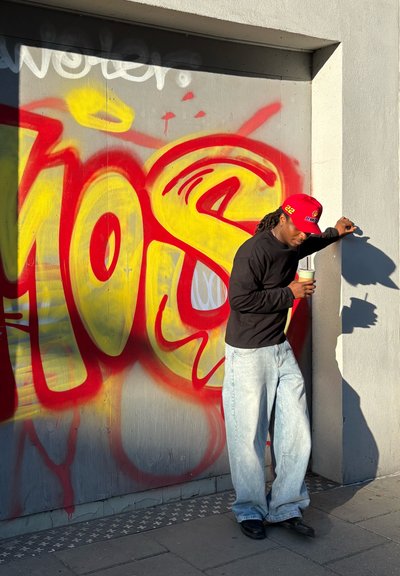 Camisa negra de manga larga, jeans anchos azules y una gorra de béisbol roja, sosteniendo una bebida frente a un fondo vibrante de graffiti amarillo y rojo.