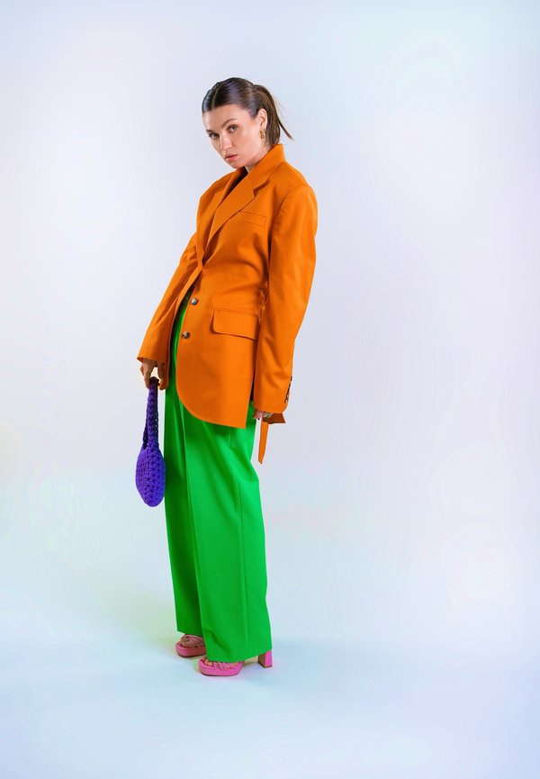 Oranger taillierter Blazer, leuchtend grüne weit geschnittene Hosen, lila gehäkelte Handtasche und pinke Absatzschuhe. Glatte Texturen und strukturierte Formen.
