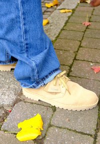 Beige suede shoes with rounded toes and textured soles, laced with tan cords, worn with blue denim jeans on a brick pavement.