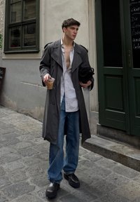 Grauer Trenchcoat, weiß gestreiftes Hemd, blaue Jeans, schwarze klobige Stiefel. Hält ein kaltes Getränk und eine schwarze Tasche. Urbaner Hintergrund mit grünem Tür.