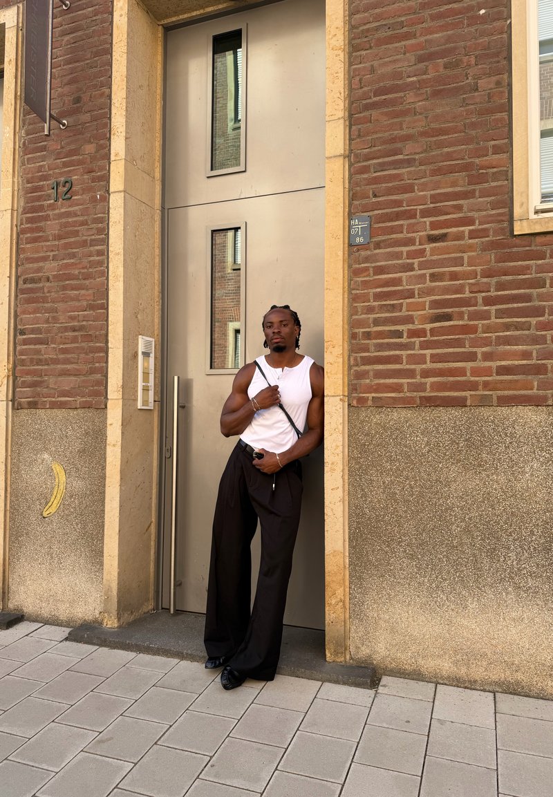 Uomo con camicia bianca senza maniche e pantaloni neri larghi appoggiato a una porta beige in un edificio di mattoni su un marciapiede piastrellato.