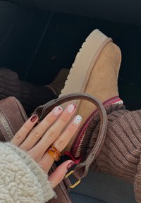 Main avec des ongles décorés marron et blancs et une bague ambre reposant sur un genou vêtu d'un pantalon en maille marron et de bottes en daim beige, tenant la poignée d'un sac marron.