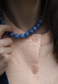 Persona que lleva un cárdigan color durazno con botones sujetando un collar azul de cuentas cerca de su cuello.