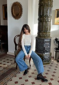 Haut blanc à manches longues côtelées, jeans larges taille haute bleu clair et bottes à talons noires. Assise sur une chaise en bois à côté d'un four.