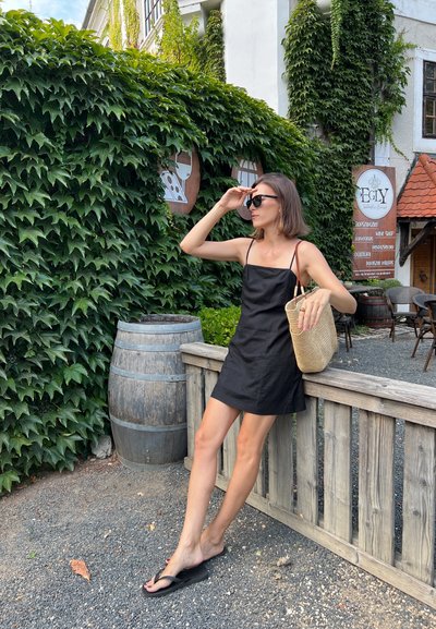 Vestido negro de lino con tirantes finos, bolso de paja texturizado y chanclas negras, posado cerca de una valla de madera con un fondo de hiedra verde.