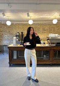 Person hält eine weiße Tasse, trägt ein schwarzes Hemd mit Knöpfen und weiße Hosen. Im Hintergrund sind eine Holztheke und eine Ziegelwand zu sehen.