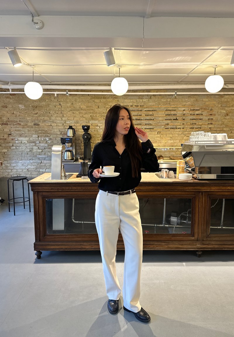 Oseba v roki drži belo skodelico, oblečena je v črno srajco z gumbi in bele hlače. V ozadju je lesen pult in opečni zid.