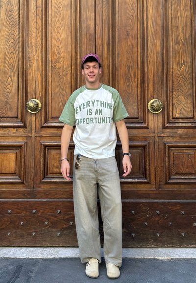 Camiseta blanca con mangas raglán verdes y texto "TODO ES UNA OPORTUNIDAD", combinada con pantalones de pierna ancha gris claro y zapatos beige.