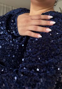 Marineblaue, mit Pailletten besetzte Kleidung mit strukturierter Oberfläche, die einen abgerundeten Ausschnitt hervorhebt. Eine Hand mit lackierten Nägeln ruht auf dem Stoff.