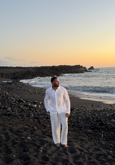 Hombre con ropa blanca de lino, de pie descalzo en una playa rocosa de arena negra al atardecer, mirando hacia el océano y el horizonte.
