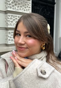 Beige ullkappa med stor knapp, lager-på-lager över en krämfärgad stickad tröja och halsduk. Guldiga iögonfallande örhängen, mjukt brunt hår, ljusrosa naglar.