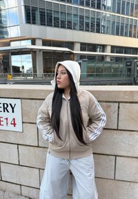 Felpa beige con zip e dettagli bianchi, dotata di cappuccio, abbinata a pantaloni corti grigi chiari. Tessuto liscio su uno sfondo in pietra texture.