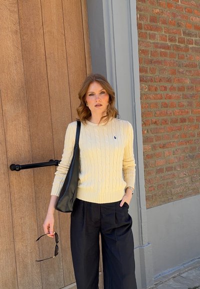 Mujer con cabello ondulado que lleva un suéter de punto trenzado crema y pantalones negros, sosteniendo gafas de sol y un bolso negro de hombro, de pie junto a una puerta de madera y una pared de ladrillos.