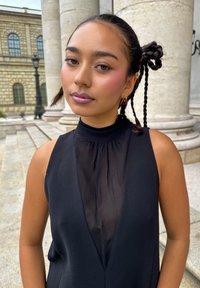 Vrouw met gevlochten donker haar dat in een bun is gestyled, gekleed in een mouwloos zwart top met doorschijnende inzet, staande in de open lucht nabij stenen kolommen en trappen.