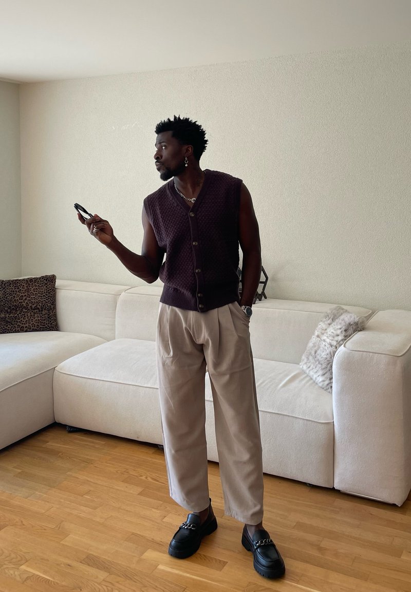 Homme portant un gilet en tricot sans manches et un pantalon beige debout près d'un canapé sectionnel blanc, tenant un smartphone et regardant sur le côté dans un salon.