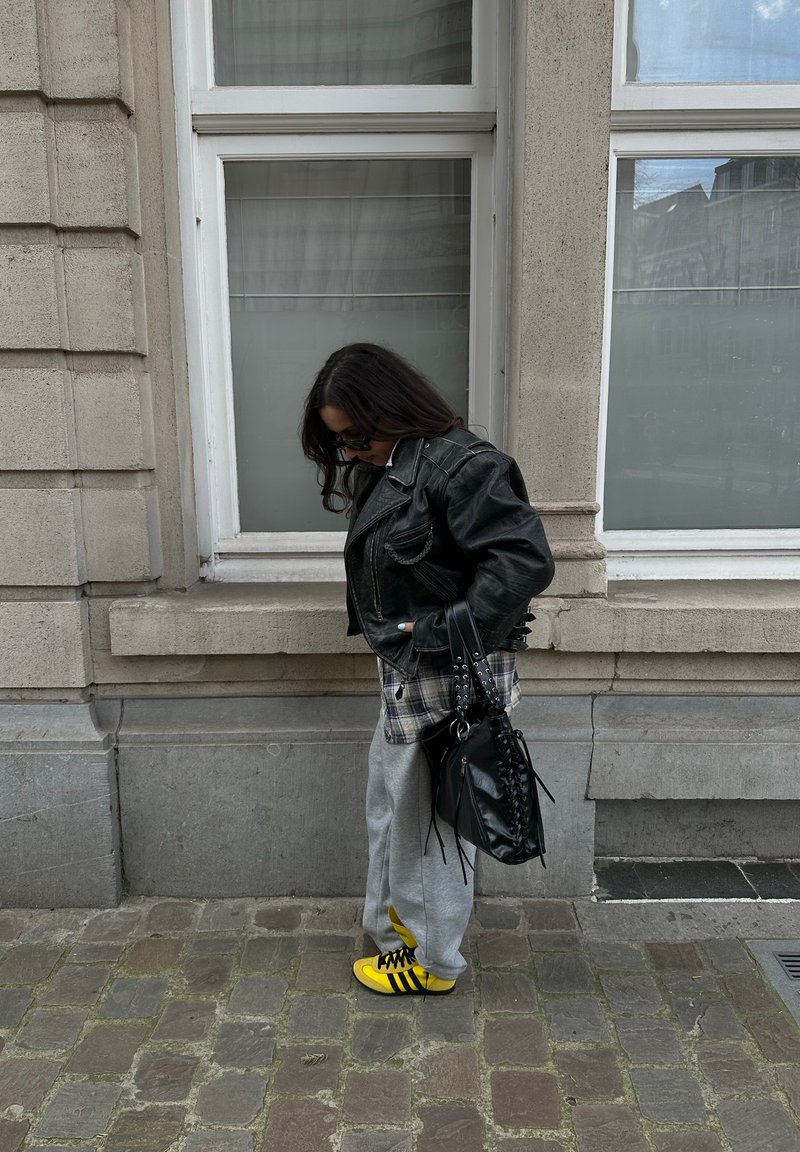 Personne portant une veste en cuir noire, un pantalon de survêtement gris et des baskets jaunes, debout dans une rue pavée à côté d’un bâtiment en pierre avec des fenêtres.