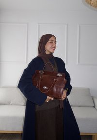 Rjava usnjena torbica s srebrnimi okovji, pravokotne oblike, dvojni ročaji in dva sprednja žepa. Model nosi moder plašč in rjave hlače.