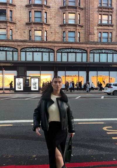 Chaqueta de cuero negra con cuello de borreguito, suéter crema, falda negra de canalé con abertura y bolso verde. Entorno urbano con escaparates.