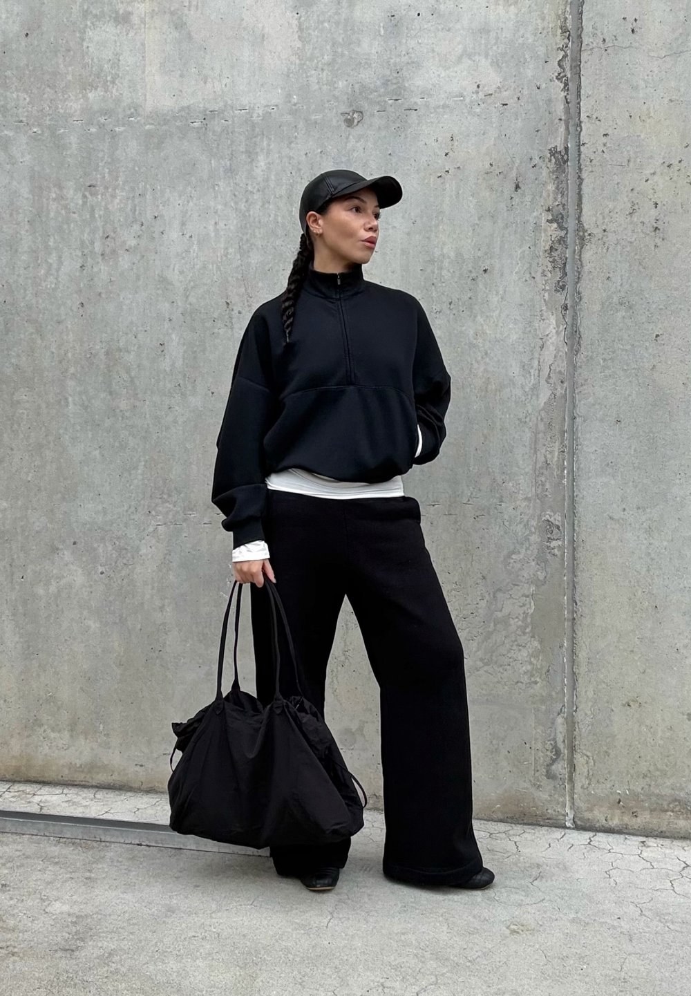 Sudadera negra con cremallera sobre una camiseta blanca, combinada con pantalones anchos negros. Un gorra negra y un bolso de tela suave y recogido en negro completan el look.