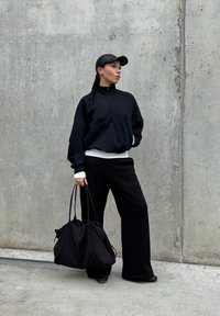 Sudadera negra con cremallera sobre una camiseta blanca, combinada con pantalones anchos negros. Un gorra negra y un bolso de tela suave y recogido en negro completan el look.