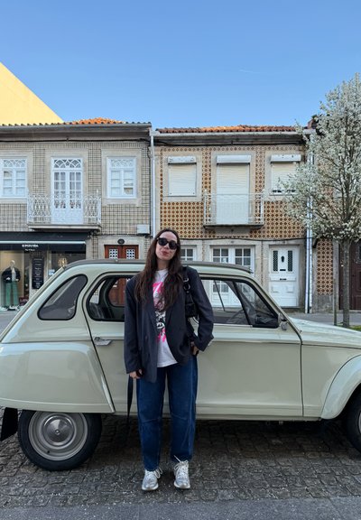 Mujer con cabello largo y oscuro, gafas de sol, ropa informal, de pie junto a un coche antiguo color crema en una calle adoquinada frente a edificios con patrones.