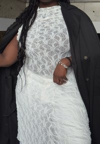 Hvid blondekjole med et tekstureret mønster, lagdelt under en sort frakke. Sølvarmbånd pryder det ene håndled og tilføjer kontrast til outfittet.