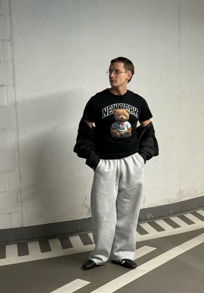 Joven con gafas, camiseta negra con un dibujo de un oso y la palabra "New York", pantalones deportivos grises y chaqueta negra, de pie en un aparcamiento cerca de marcas en la pared.